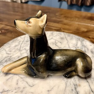 May include: A ceramic dog figurine in a reclining pose. The dog has a black body with gold accents and a tan head and paws. The figurine is resting on a marble-patterned surface.