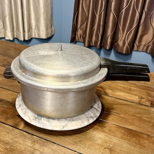 May include: A vintage aluminum pressure cooker with a black handle and lid. The pressure cooker sits on a round, white and gray marble base. The kitchenware is on a wooden surface.