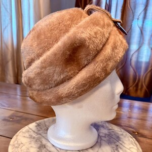 May include: A vintage-style, tan-colored faux fur hat. The hat is displayed on a white mannequin head, showcasing its textured, plush material and curved design. A dark brown clasp secures the hat's strap. The hat sits on a marble-patterned surface.
