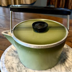 Può includere: Un bollitore verde oliva con bordo in metallo argentato e manico in plastica nera. Il coperchio presenta una manopola centrale nera con un piccolo logo circolare in rilievo. Il bollitore ha un beccuccio integrato sul lato sinistro.