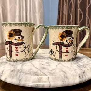 May include: Two ceramic mugs with a snowman design. The mugs have a cream-coloured base with a green rim and handle. Each mug features a snowman with a black hat, orange nose, and a red and green scarf. The snowman has three red heart-shaped buttons.