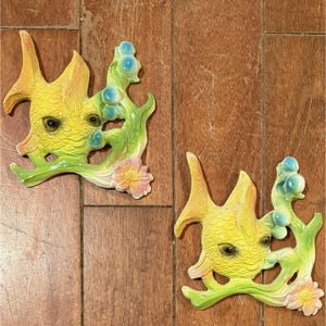 May include: Two ceramic fish wall decorations. The fish are yellow with orange accents, black eyes, and green and blue seaweed. Each fish has a pink and yellow flower.