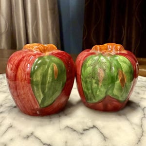 May include: A pair of ceramic apple-shaped salt and pepper shakers. The shakers are red with green leaf accents and orange stems. They are on a marble surface.