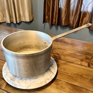 May include: A silver-colored saucepan with a long handle, resting on a white and gray marble trivet. The pot is empty and sits on a wooden table. The handle has a hole at the end for hanging.
