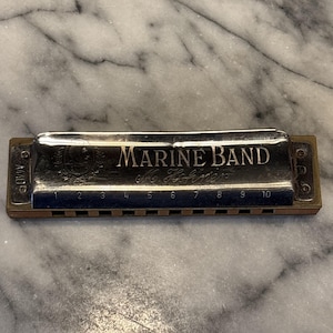 May include: A silver Marine Band harmonica with the text "MARINE BAND" and "M. Hohner" engraved on the top. The harmonica has ten numbered holes and is made of metal and wood.
