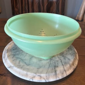 Puede incluir: Un colador de plástico de color verde menta claro con un borde ancho y redondeado y una serie de pequeños orificios de drenaje circulares dispuestos en un patrón triangular en la base. El utensilio de cocina tiene un acabado liso y mate.