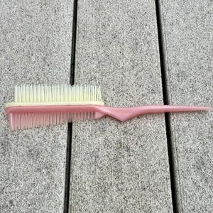 May include: A pink hairbrush and comb combination. The brush has a light beige bristle section and a pink handle. The comb section is also pink. The brush is approximately 9 inches long.