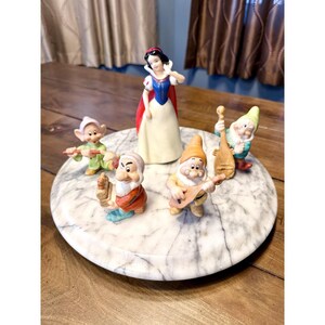 以下が含まれることがあります： 白雪姫と七人の小人をフィーチャーしたディズニーのフィギュアのコレクション。フィギュアは、丸い大理石模様の表面に配置されています。白雪姫は青と赤のドレスを着ており、小人たちは様々な衣装を着ています。