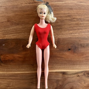 May include: Vintage doll with blonde hair styled in a ponytail with a black bow. The doll is wearing a red swimsuit. The doll's skin is a pale pink color. The doll is standing on a wooden surface.