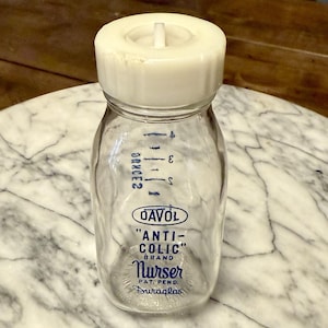 May include: Clear glass baby bottle with a white plastic cap. The bottle has blue measurement markings and the text "DAVOL ANTI-COLIC BRAND Nurser". The bottle is on a marble surface.
