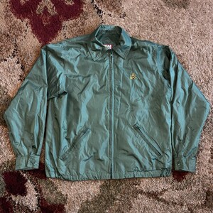 May include: A sage green jacket with a full-length zipper, two front pockets, and a collared neckline. The jacket has long sleeves with buttoned cuffs and a small embroidered emblem on the chest. The jacket is made of a lightweight, possibly nylon, material.