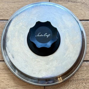 May include: A circular stainless steel pot lid with a polished metallic finish and a black, star-shaped heat-resistant knob in the center. The knob features the white script text Lustre Craft printed on its top surface, providing a clear brand identifier.