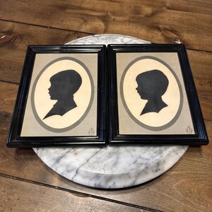 May include: Two rectangular black frames containing identical silhouette portraits of a child in profile. The silhouettes are black against a cream-colored oval background, which is set within a textured gray rectangular border inside each frame.