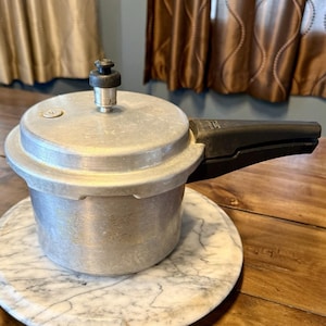 May include: A silver pressure cooker with a black handle and a pressure release valve. The cooker sits on a white and gray marble-patterned surface. The kitchen appliance is designed for cooking food quickly.