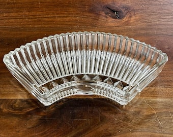 Puede incluir: Un plato de vidrio transparente en forma de abanico con detalles acanalados verticales. El plato tiene un borde festoneado y un patrón geométrico en la base. El plato se utiliza probablemente para servir aperitivos o exhibir objetos decorativos.