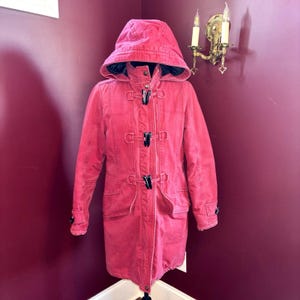May include: A red hooded coat with a toggle closure. The coat has a long length, two front pockets, and black toggles. The coat is displayed on a mannequin against a burgundy wall.
