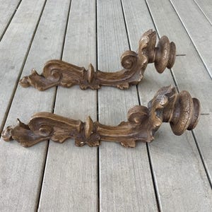 Puede incluir: Dos candelabros de pared a juego con un acabado en tono madera marrón. Cada pieza presenta un diseño alargado y curvo con volutas, detalles tallados intrincados y una plataforma circular escalonada en el extremo para sostener una vela.