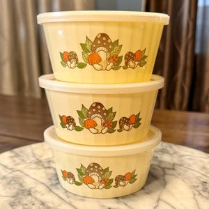 May include: Three yellow plastic bowls with lids stacked on top of each other. Each bowl has a repeating pattern of mushrooms in brown, orange, and white with green leaves. The bowls are on a marble surface.