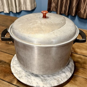 May include: A vintage aluminum pot with a lid and black handles. The pot has a capacity of 6 quarts and sits on a marble-patterned trivet. The lid has a red knob.