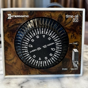 May include: A square Intermatic time-all XV timer with a wood grain face. The timer has a black dial with white numbers and markings for AM and PM. The timer has a push on-off switch and a time setting lever.