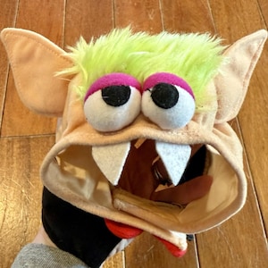 May include: A monster hat with large, cartoonish eyes, white felt teeth, and a red tongue. The hat has a tan base with lime green fur and large tan ears. The hat is being held by a person.