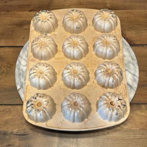 Puede incluir: Un molde para bizcocho bundt vintage, de color blanquecino, con 12 moldes individuales. El molde tiene forma rectangular con bordes redondeados. Cada molde tiene forma de flor. El molde tiene un aspecto ligeramente desgastado.