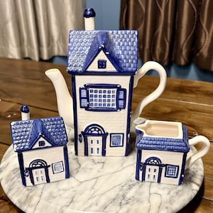 May include: A white and blue ceramic tea set in the shape of houses. The set includes a teapot, a creamer, and a sugar bowl. The roofs are blue, and the walls are white with blue trim. The teapot has a tall chimney.