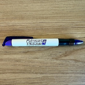 May include: A Pulmicort Turbuhaler inhaler, featuring a white body with blue and black accents. The text "Pulmicort 200 mcg Turbuhaler (budesonide inhalation powder)" is printed on the inhaler. The inhaler is on a wooden surface.