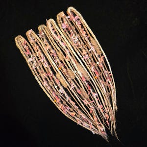May include: A set of five long, light brown hair extensions featuring intricate wraps of pink and white fabric strips, thin twine, and small wooden beads. Each extension is constructed with a looped top and tapers toward the bottom ends.