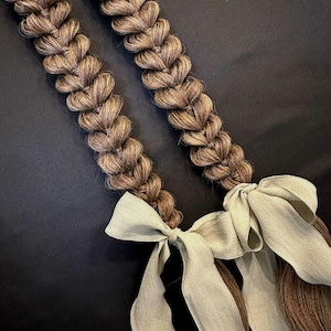 May include: Two long, braided hair extensions in shades of brown, secured with light green fabric bows. The braids are thick and detailed, with a textured appearance. The background is a solid black.