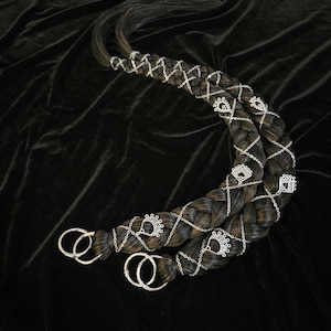 May include: Two dark brown braided hair extensions with silver-colored decorative chains and charms. The braids are adorned with small, ornate silver accents and connected by silver chains. The ends of the braids have silver rings.