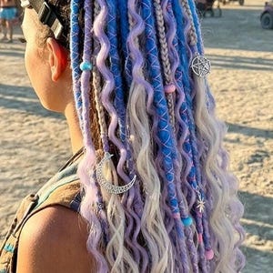 May include: A set of hair extensions featuring a mix of blue, lavender, and white wavy strands styled in a high bun. The hair includes braided sections, colorful beads, and silver-toned metal charms shaped like a crescent moon and a pentagram.