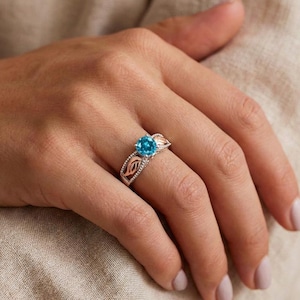 May include: A silver ring with a large, round, bright blue gemstone. The ring has a decorative design with rose gold accents on the sides. The ring is on a person's finger.