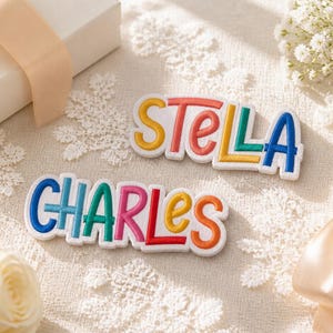 Pode incluir: Dois emblemas de tecido branco com nomes bordados em letras de bloco coloridas. O emblema superior exibe o nome STELLA em amarelo, vermelho, azul-petróleo e azul. O emblema inferior exibe o nome CHARLES em azul, azul-petróleo, rosa, amarelo e laranja.