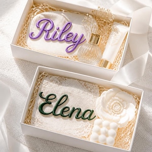 May include: Two white gift boxes containing personalized items. The top box features a pouch with the purple embroidered name Riley, a glass perfume bottle, and a tube. The bottom box holds a lace square with the green embroidered name Elena, a white flower-shaped candle, and a white square bubble candle.