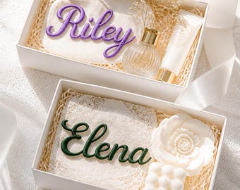 Könnte beinhalten: Zwei weiße Geschenkboxen mit personalisierten Artikeln. Die obere Box enthält ein Täschchen mit dem violett gestickten Namen Riley, einen Glasparfümflakon und eine Tube. Die untere Box enthält ein Spitzenquadrat mit dem grün gestickten Namen Elena, eine weiße blumenförmige Kerze und eine weiße quadratische Bubble-Kerze.