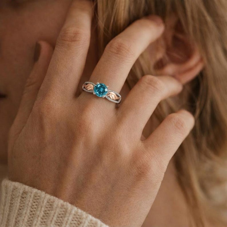 May include: A silver ring with a round, bright blue gemstone. The ring features rose gold leaf accents on the sides and small clear stones. The ring is on a person's finger.