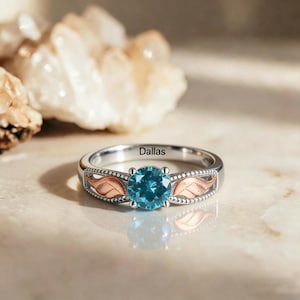 May include: A silver ring with a large, round, blue gemstone. The ring has rose gold-colored leaf accents on each side of the gemstone and the word "Dallas" engraved on the band.