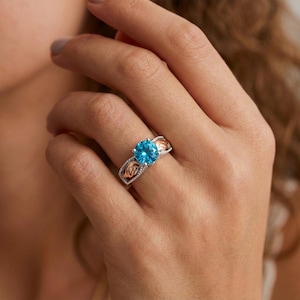 May include: A silver ring with a large, round, bright blue gemstone. The ring has rose gold accents on the sides with a decorative pattern. The ring is on a person's finger.