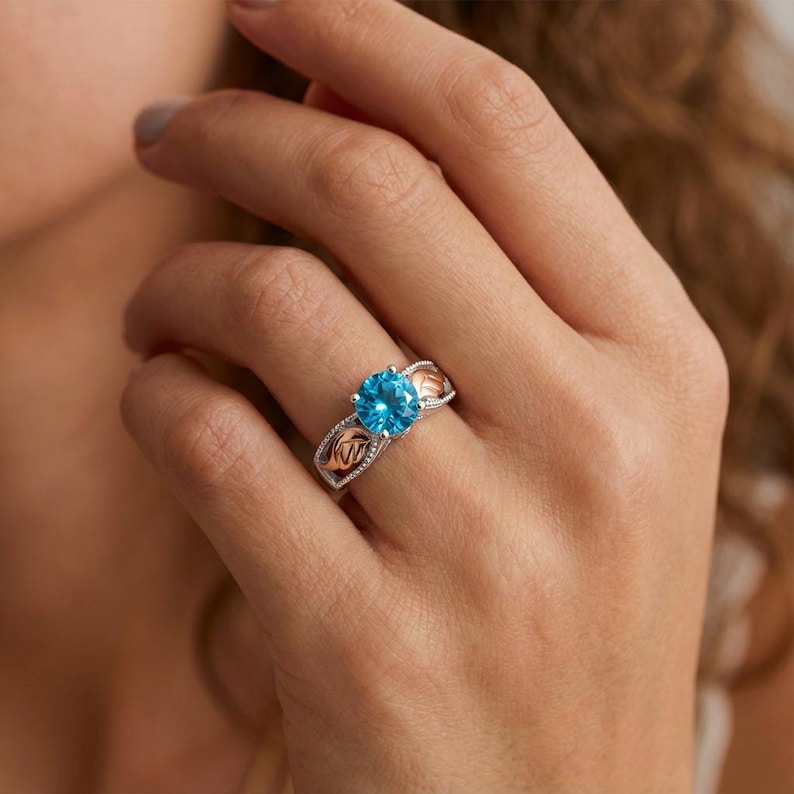 May include: A silver ring with a large, round, bright blue gemstone. The ring has rose gold accents on the sides with a decorative pattern. The ring is on a person's finger.