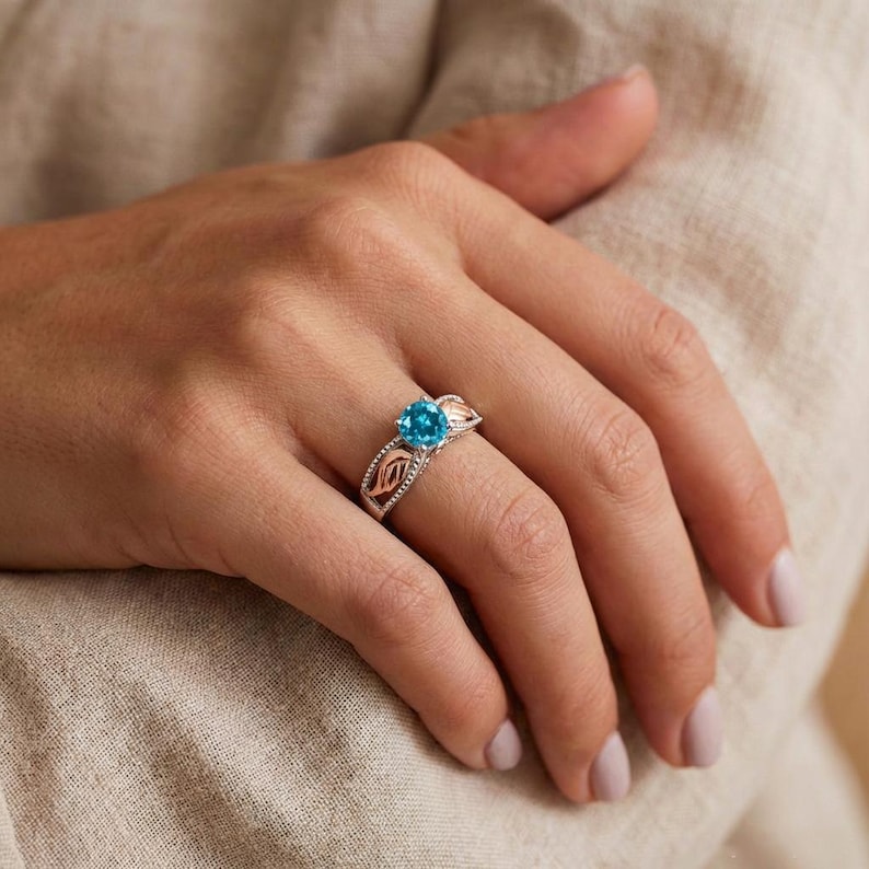 May include: A silver ring with a large, round, bright blue gemstone. The ring has a decorative design with rose gold accents on the sides. The ring is on a person's finger.