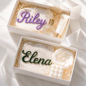 K&ouml;nnte beinhalten: Zwei wei&szlig;e Geschenkboxen mit personalisierten Artikeln. Die obere Box enth&auml;lt ein T&auml;schchen mit dem violett gestickten Namen Riley, einen Glasparf&uuml;mflakon und eine Tube. Die untere Box enth&auml;lt ein Spitzenquadrat mit dem gr&uuml;n gestickten Namen Elena, eine wei&szlig;e blumenf&ouml;rmige Kerze und eine wei&szlig;e quadratische Bubble-Kerze.