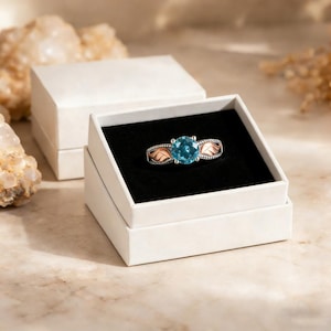 May include: A silver ring with a large, round, light blue gemstone is displayed in a white box with a black interior. The ring has rose gold-colored accents on the sides. The box is open, and another closed box is in the background.