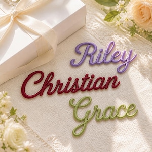 K&ouml;nnte beinhalten: Drei gestickte Namensaufn&auml;her in Schreibschrift. Der oberste Aufn&auml;her zeigt den Namen Riley in Lila, der mittlere Christian in Dunkelrot und der untere Grace in Hellgr&uuml;n. Die Aufn&auml;her sind auf einer strukturierten Stoffoberfl&auml;che angeordnet.