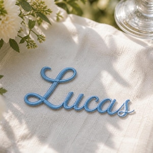 K&ouml;nnte beinhalten: Ein hellblauer, gestickter Namensaufn&auml;her mit dem Wort Lucas in einer kursiven Schriftart. Die Stickerei ist auf eine strukturierte, cremewei&szlig;e Stoffoberfl&auml;che aufgebracht und zeigt ein sauberes, erhabenes Fadendesign in einheitlichem Blau.