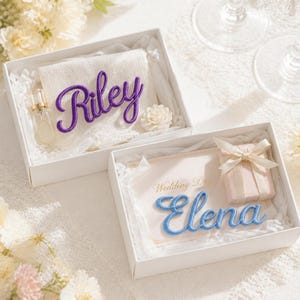 K&ouml;nnte beinhalten: Zwei wei&szlig;e Geschenkboxen mit personalisierten Artikeln. Eine Box enth&auml;lt ein wei&szlig;es Handtuch mit dem violetten Namen Riley und ein kleines Parf&uuml;mfl&auml;schchen. Die andere Box enth&auml;lt eine Karte mit dem blauen Namen Elena und ein kleines Geschenk.