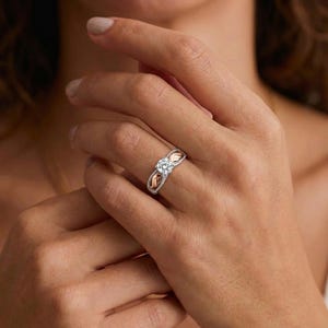 Pode incluir: Um anel de prata com uma pedra preciosa central, redonda e transparente. O anel apresenta um design decorativo com detalhes dourados nas laterais. O anel é usado num dedo.