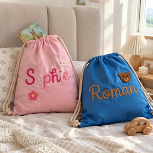 Pode incluir: Uma bolsa de cordão rosa bordada com o nome Sophie e duas pequenas flores aplicadas, e uma bolsa de cordão azul bordada com o nome Roman e um aplique de urso castanho, ambas com cordões grossos de cor creme.