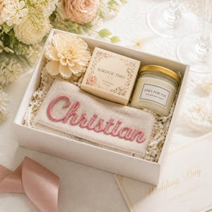 K&ouml;nnte beinhalten: Eine wei&szlig;e Geschenkbox mit einem beigen Stoffbeutel, der mit dem Namen Christian in rosa Garn bestickt ist, einer quadratischen Schachtel mit der Aufschrift Romantic Times, einer Glas-Duftkerze mit dem Etikett Only For You und einer cremefarbenen Kunstblume.