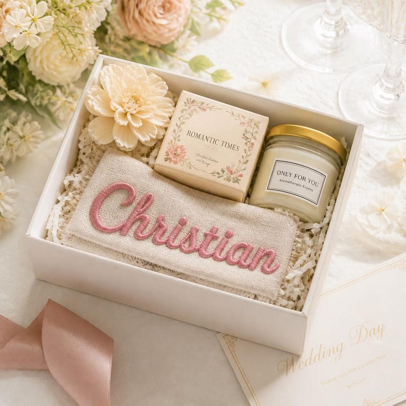 K&ouml;nnte beinhalten: Eine wei&szlig;e Geschenkbox mit einem beigen Stoffbeutel, der mit dem Namen Christian in rosa Garn bestickt ist, einer quadratischen Schachtel mit der Aufschrift Romantic Times, einer Glas-Duftkerze mit dem Etikett Only For You und einer cremefarbenen Kunstblume.